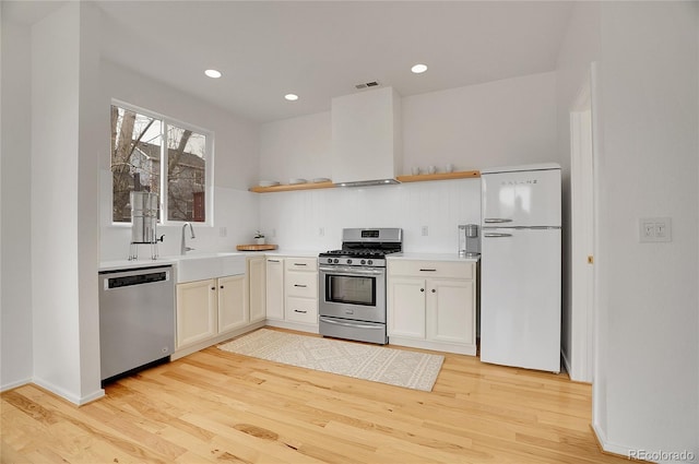 kitchen featuring light wood-style floors, stainless steel appliances, light countertops, wall chimney range hood, and open shelves