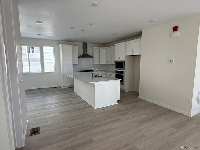 kitchen with wall chimney range hood, sink, appliances with stainless steel finishes, a kitchen island with sink, and white cabinetry