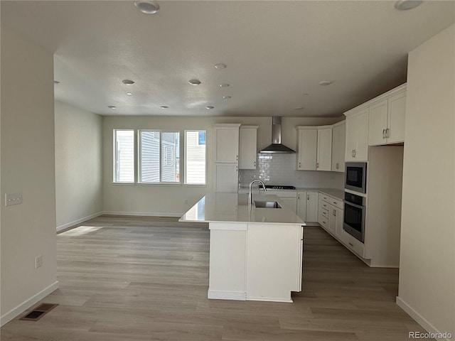 kitchen with sink, black appliances, a center island with sink, wall chimney range hood, and white cabinets