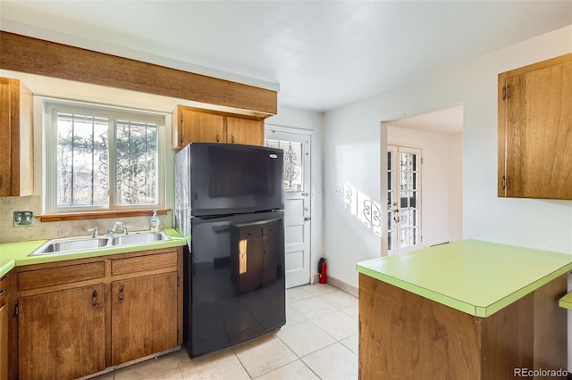 kitchen with brown cabinetry, light tile patterned flooring, freestanding refrigerator, a sink, and light countertops