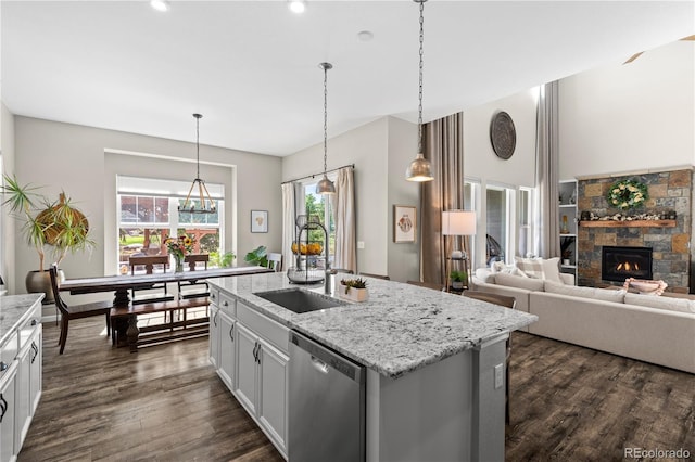 kitchen with dark wood-style flooring, light stone counters, a stone fireplace, an island with sink, and stainless steel dishwasher