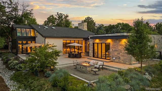 back of house at dusk featuring stucco siding, stone siding, a fire pit, a chimney, and a patio area