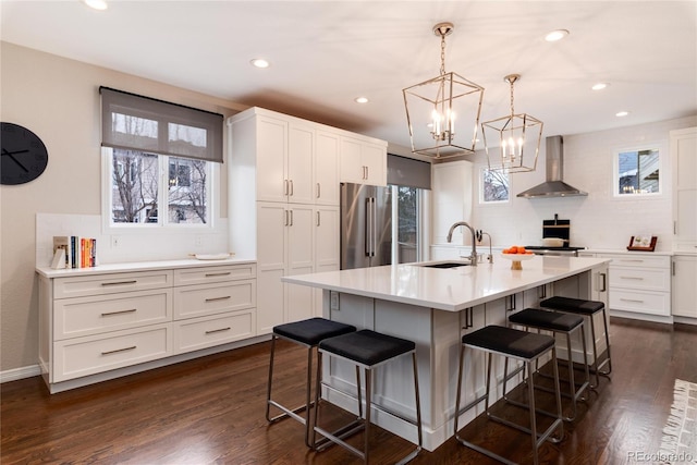 kitchen featuring a center island with sink, high end refrigerator, light countertops, a sink, and wall chimney exhaust hood