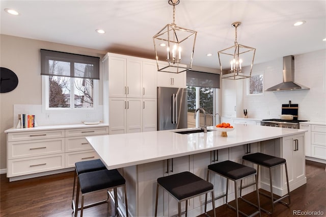 kitchen featuring freestanding refrigerator, a kitchen island with sink, light countertops, wall chimney range hood, and a sink