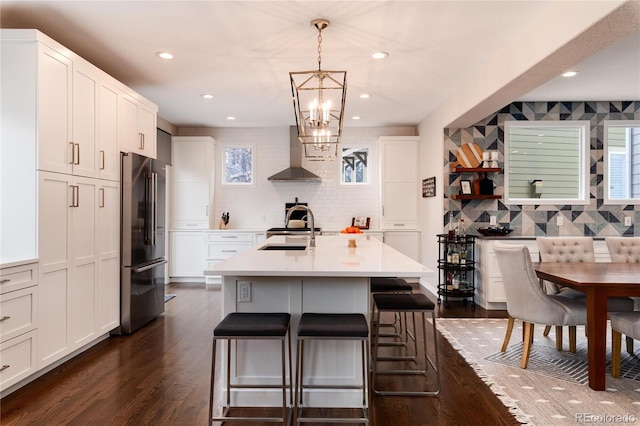 kitchen featuring decorative light fixtures, light countertops, high quality fridge, an island with sink, and wall chimney exhaust hood