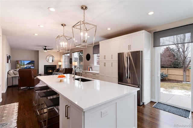 kitchen with light countertops, freestanding refrigerator, white cabinets, a kitchen island with sink, and a warm lit fireplace