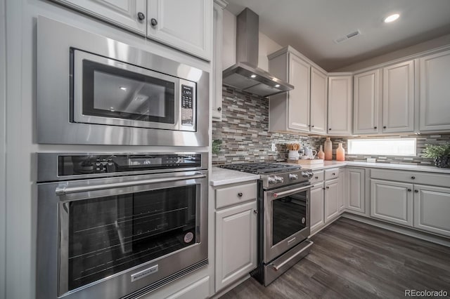 kitchen with wall chimney range hood, appliances with stainless steel finishes, backsplash, dark hardwood / wood-style floors, and white cabinets