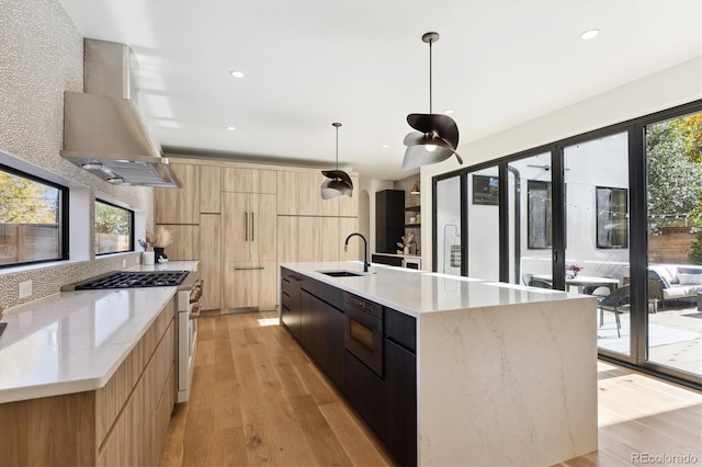 kitchen with a large island, sink, hanging light fixtures, wall chimney range hood, and light wood-type flooring