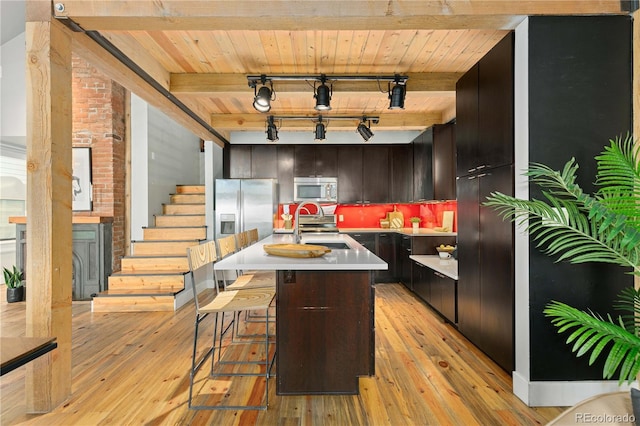 kitchen featuring a kitchen bar, an island with sink, stainless steel appliances, track lighting, and beamed ceiling