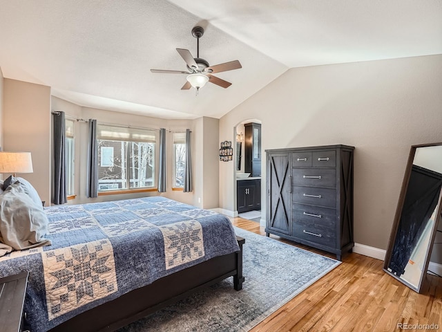 bedroom with arched walkways, baseboards, vaulted ceiling, light wood finished floors, and ensuite bath