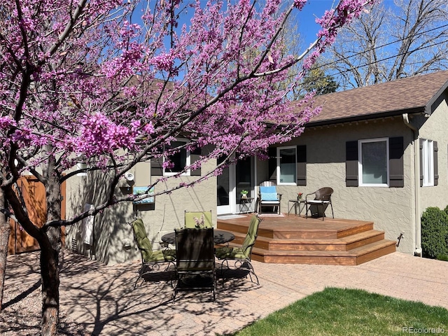 rear view of property featuring stucco siding, a patio, a wooden deck, and a shingled roof