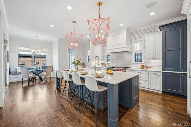 kitchen with premium range hood, decorative light fixtures, an island with sink, white cabinets, and crown molding