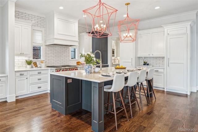 kitchen featuring premium range hood, backsplash, white cabinetry, a kitchen island with sink, and stainless steel gas cooktop
