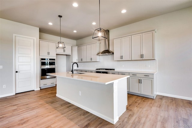 kitchen with sink, hanging light fixtures, a kitchen island with sink, stainless steel appliances, and wall chimney range hood