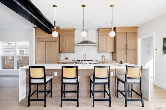 kitchen with wall chimney range hood, light hardwood / wood-style floors, an island with sink, decorative light fixtures, and tasteful backsplash