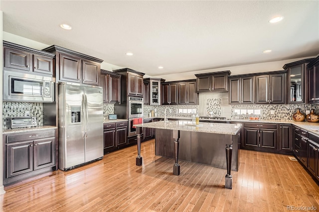 kitchen featuring a kitchen island with sink, dark brown cabinetry, appliances with stainless steel finishes, light wood finished floors, and a kitchen bar