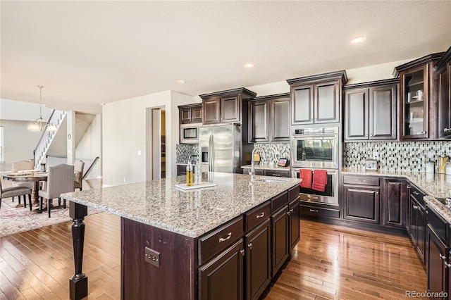 kitchen featuring stainless steel appliances, a sink, backsplash, an island with sink, and dark wood finished floors