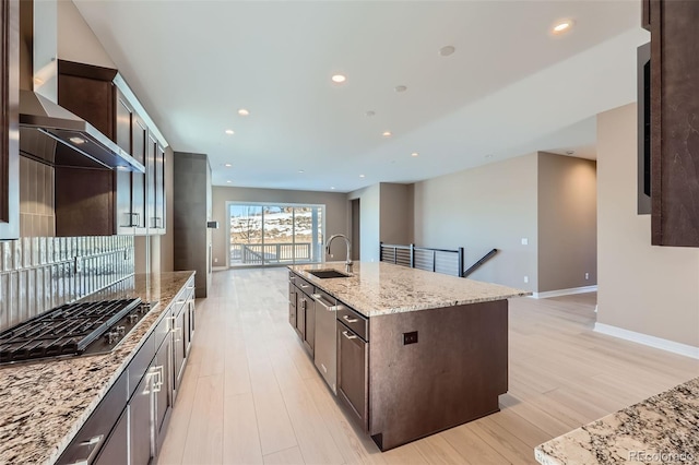 kitchen with a center island with sink, stainless steel gas stovetop, light stone counters, wall chimney exhaust hood, and sink