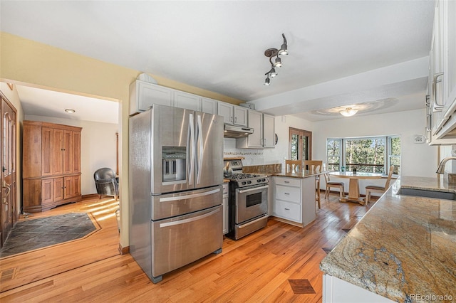 kitchen featuring light wood finished floors, appliances with stainless steel finishes, a peninsula, under cabinet range hood, and a sink