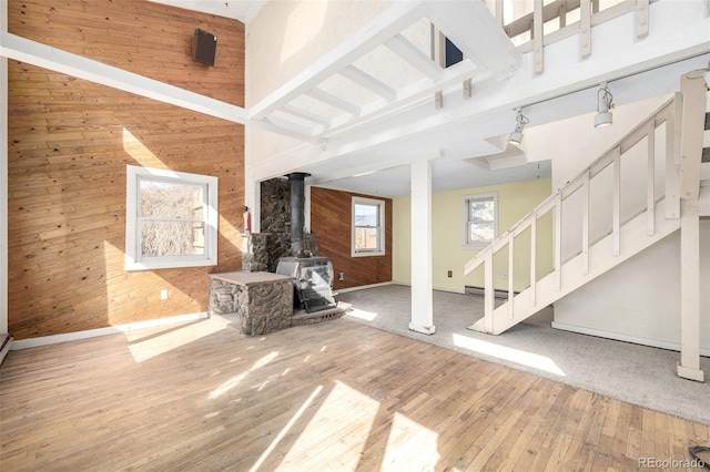 unfurnished living room with hardwood / wood-style flooring, a baseboard radiator, stairway, a wood stove, and wood walls