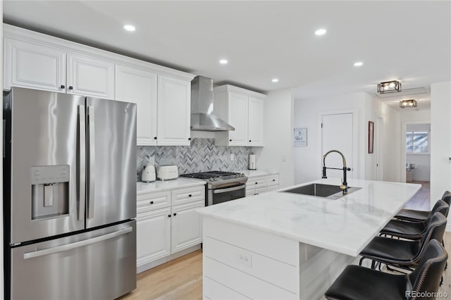 kitchen with sink, a kitchen island with sink, white cabinetry, stainless steel appliances, and wall chimney exhaust hood