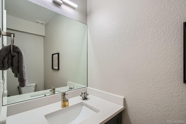 bathroom with visible vents, toilet, vanity, and a textured wall