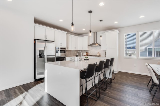 kitchen with white cabinetry, stainless steel appliances, a center island with sink, decorative light fixtures, and wall chimney exhaust hood