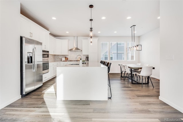 kitchen featuring wall chimney exhaust hood, white cabinetry, decorative light fixtures, an island with sink, and stainless steel appliances