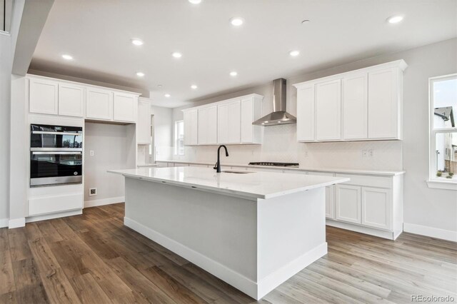 kitchen featuring stainless steel double oven, white cabinets, a kitchen island with sink, a sink, and wall chimney range hood