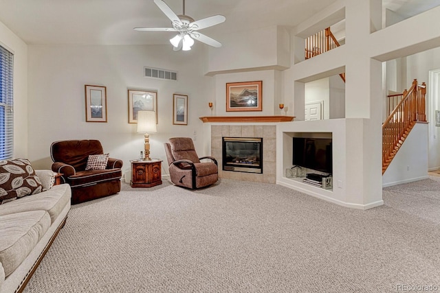 carpeted living room featuring visible vents, stairs, high vaulted ceiling, a tile fireplace, and a ceiling fan