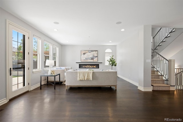 living room featuring stairway, a warm lit fireplace, dark wood-style floors, baseboards, and recessed lighting