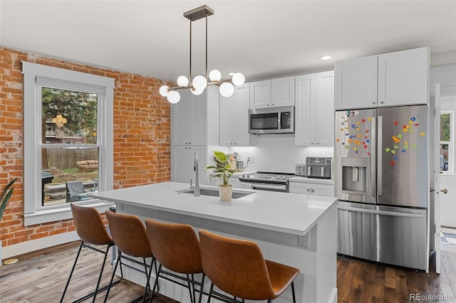 kitchen with brick wall, appliances with stainless steel finishes, white cabinetry, dark wood-type flooring, and a center island with sink