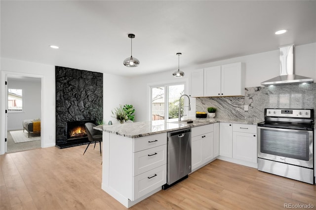 kitchen with wall chimney exhaust hood, a peninsula, a sink, a stone fireplace, and appliances with stainless steel finishes