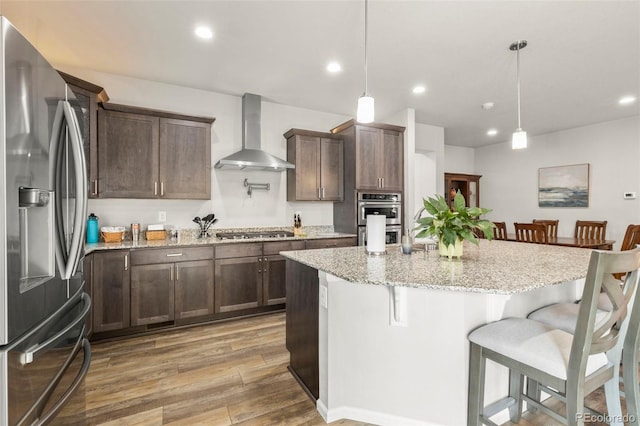 kitchen featuring stainless steel appliances, wall chimney exhaust hood, light wood-style floors, a breakfast bar area, and dark brown cabinets