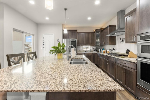 kitchen featuring stainless steel appliances, wall chimney range hood, a sink, dark brown cabinets, and a large island