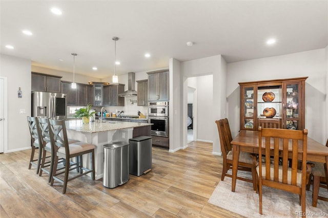 kitchen featuring stainless steel appliances, wall chimney range hood, light wood-type flooring, a breakfast bar, and recessed lighting