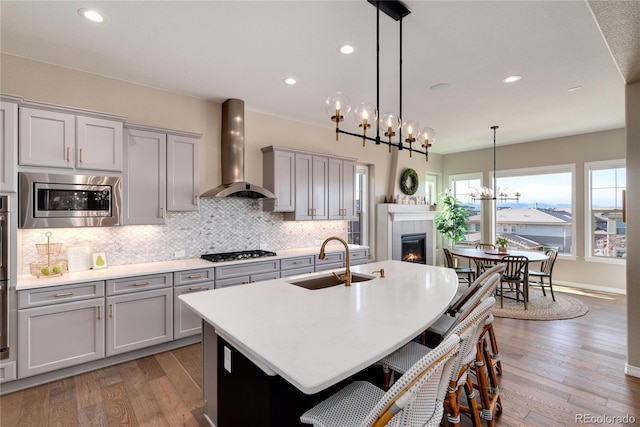 kitchen with an island with sink, stainless steel appliances, gray cabinetry, wall chimney range hood, and a sink