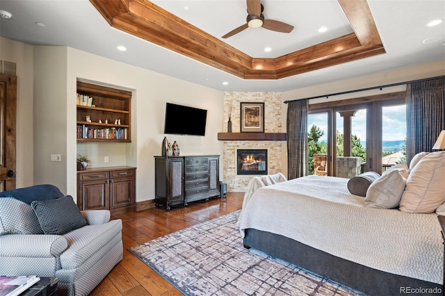 bedroom with a stone fireplace, dark hardwood / wood-style floors, ceiling fan, access to exterior, and a tray ceiling