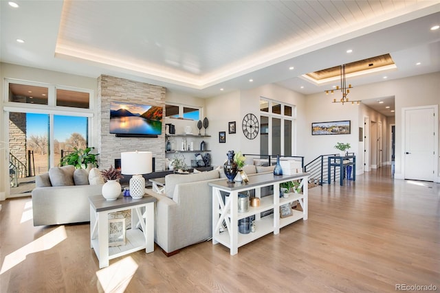 living area featuring a tray ceiling, a stone fireplace, recessed lighting, light wood-style flooring, and a notable chandelier