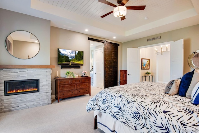 bedroom with light carpet, a glass covered fireplace, recessed lighting, a barn door, and a raised ceiling