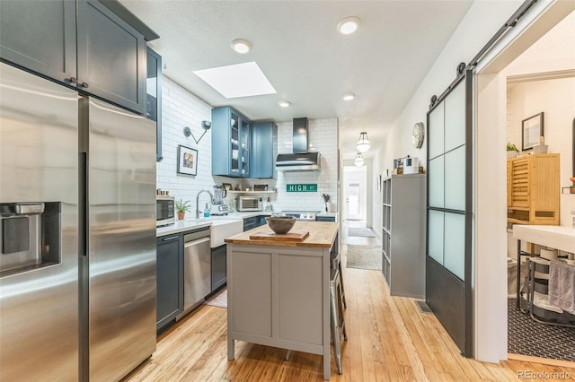 kitchen with a barn door, stainless steel appliances, wood counters, wall chimney range hood, and light wood-type flooring