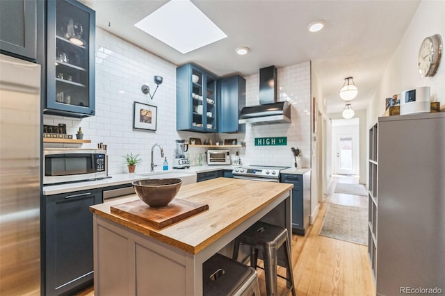 kitchen with light wood-style flooring, blue cabinets, stainless steel appliances, butcher block counters, and wall chimney range hood