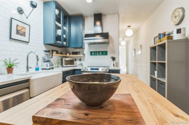 kitchen featuring blue cabinetry, decorative backsplash, glass insert cabinets, wall chimney range hood, and range