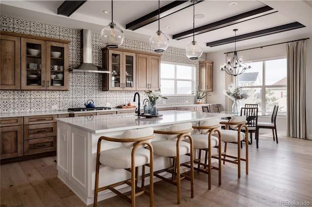 kitchen with wall chimney range hood, beam ceiling, a kitchen island with sink, hardwood / wood-style floors, and backsplash