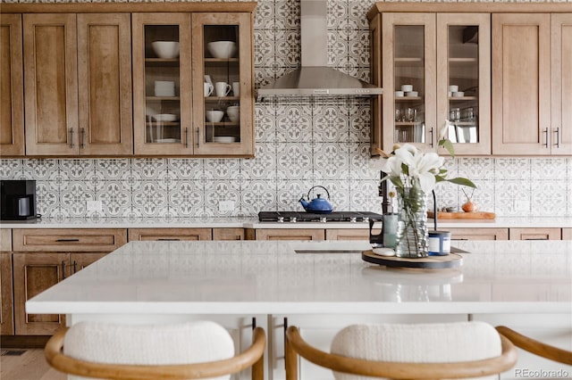 kitchen with brown cabinetry, decorative backsplash, glass insert cabinets, light countertops, and wall chimney exhaust hood