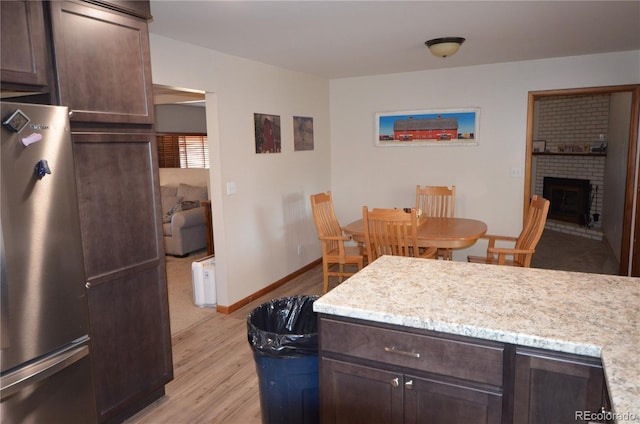 kitchen with dark brown cabinetry, baseboards, freestanding refrigerator, light wood-type flooring, and a brick fireplace