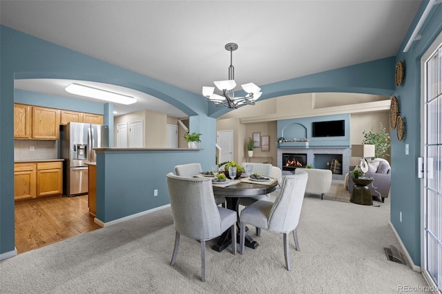 dining room featuring baseboards, a lit fireplace, light carpet, an inviting chandelier, and arched walkways