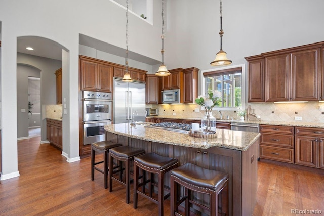 6299 Ellingwood Point Pl, Castle Rock, CO 80108 - kitchen featuring appliances with stainless steel finishes, dark wood finished floors, a kitchen island with sink, light stone counters, and a towering ceiling