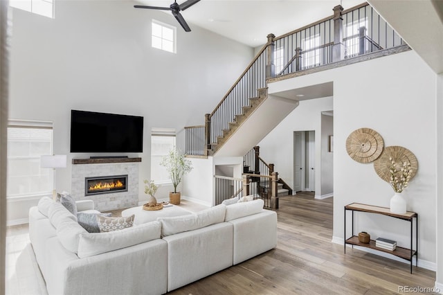 living area with ceiling fan, a tiled fireplace, wood finished floors, a towering ceiling, and stairway