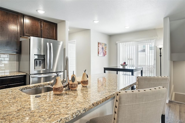 kitchen with light stone countertops, tasteful backsplash, dark brown cabinets, stainless steel refrigerator with ice dispenser, and wood finished floors